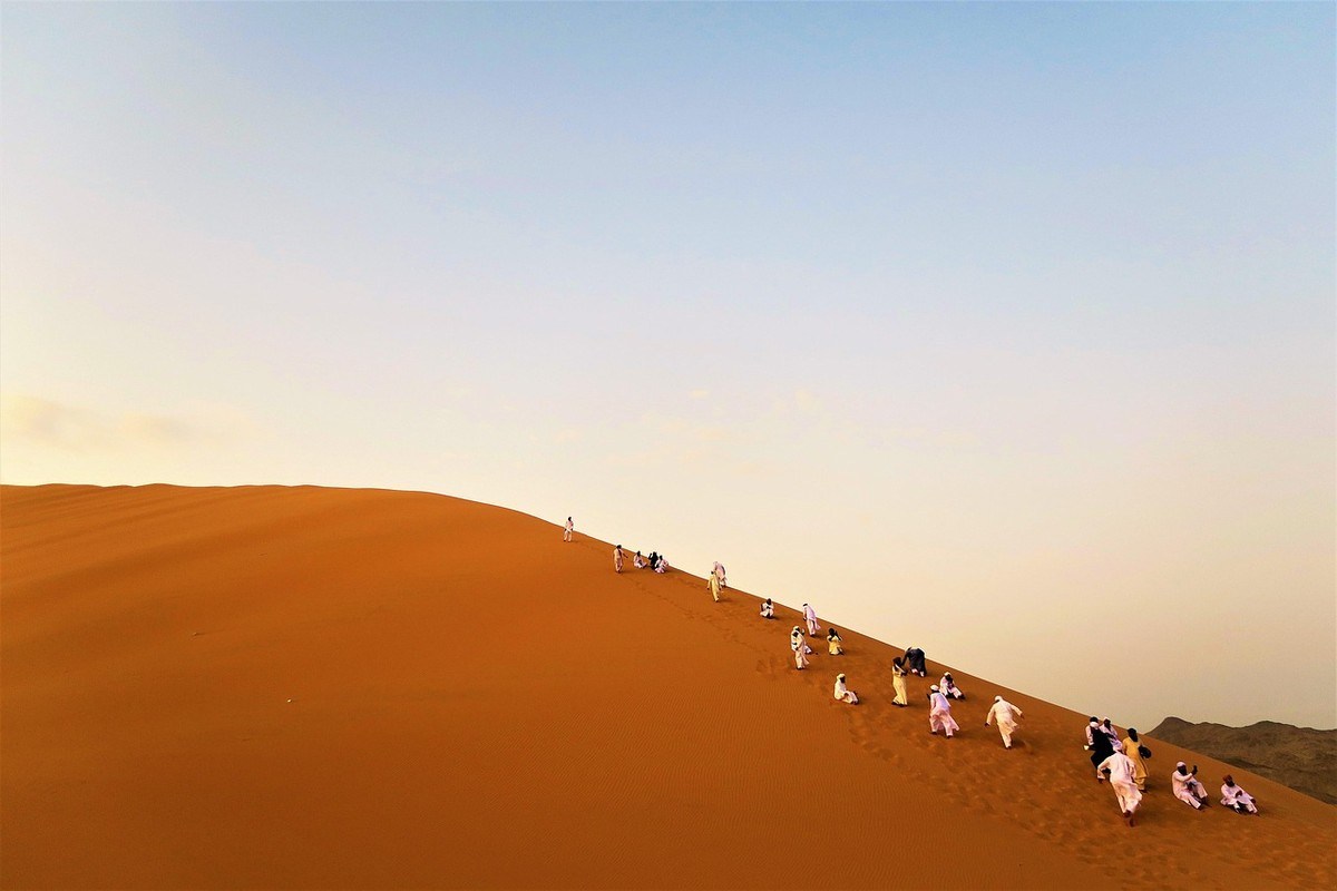 File de trekkers progressant sur une dune de sable dans un désert aride au coucher de soleil File de trekkers progressant sur une dune de sable dans un désert aride au coucher de soleil